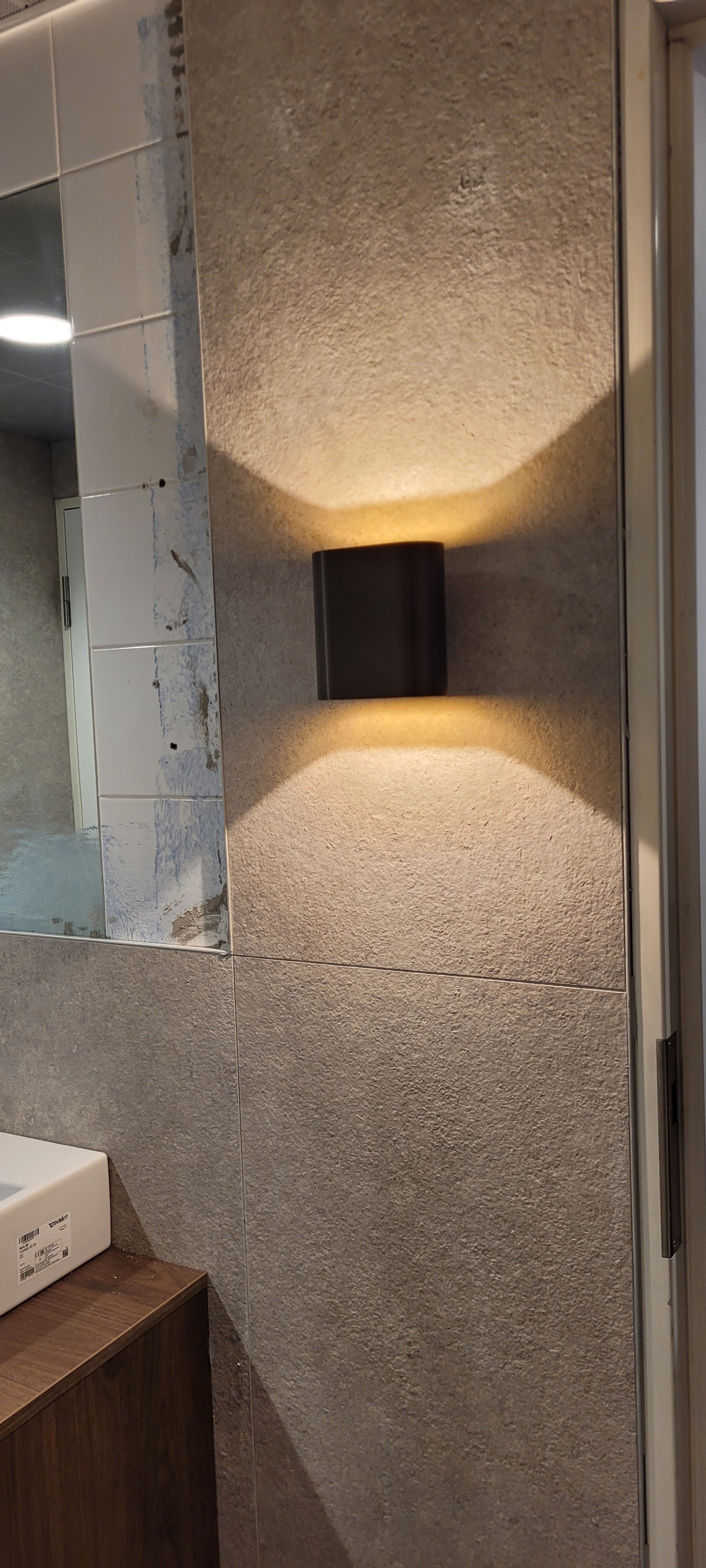 Zwarte wandlamp met licht naar boven en beneden op een betegelde badkamerwand naast een spiegel